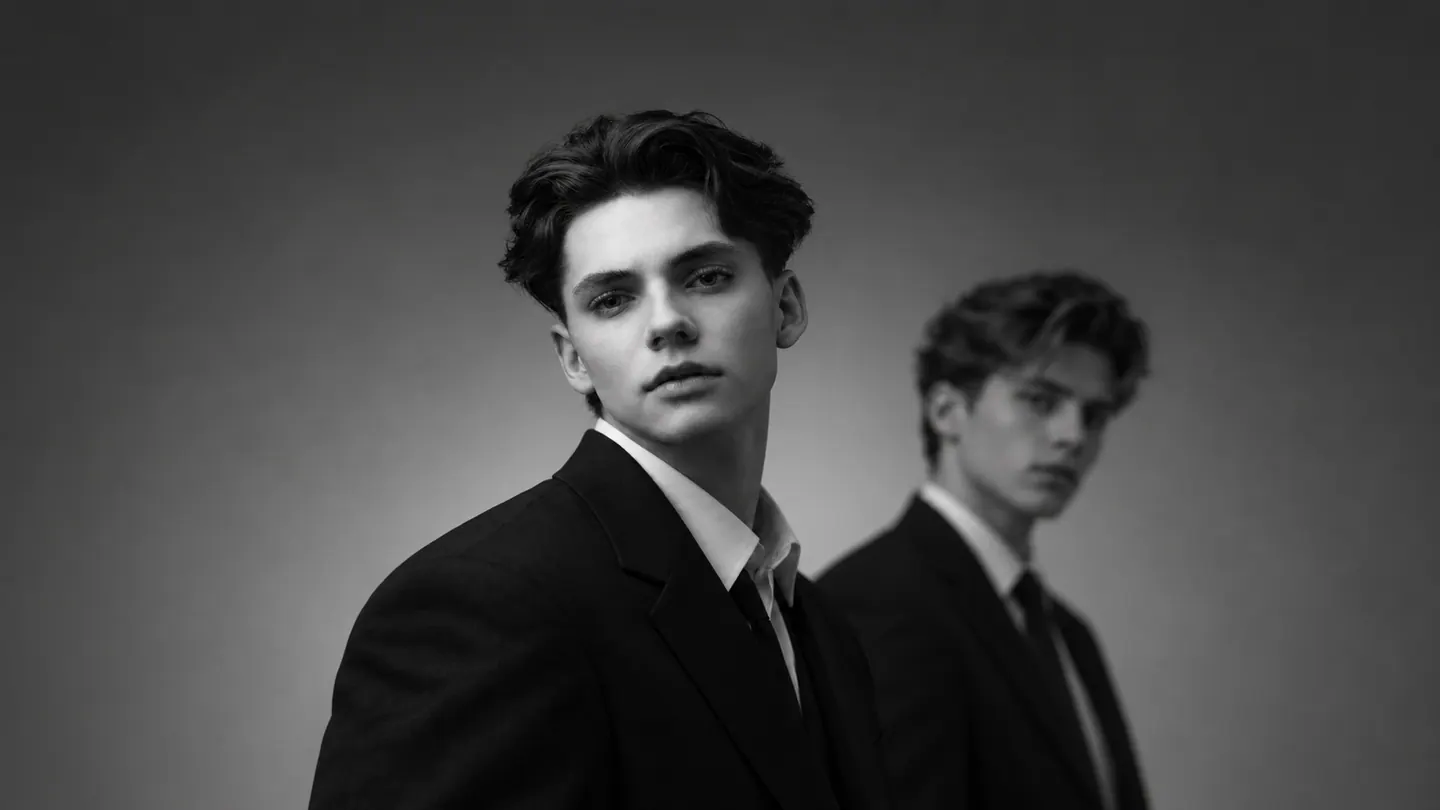 Retrato editorial en blanco y negro de dos hombres jóvenes con trajes oscuros, uno nítido en primer plano y otro desenfocado al fondo