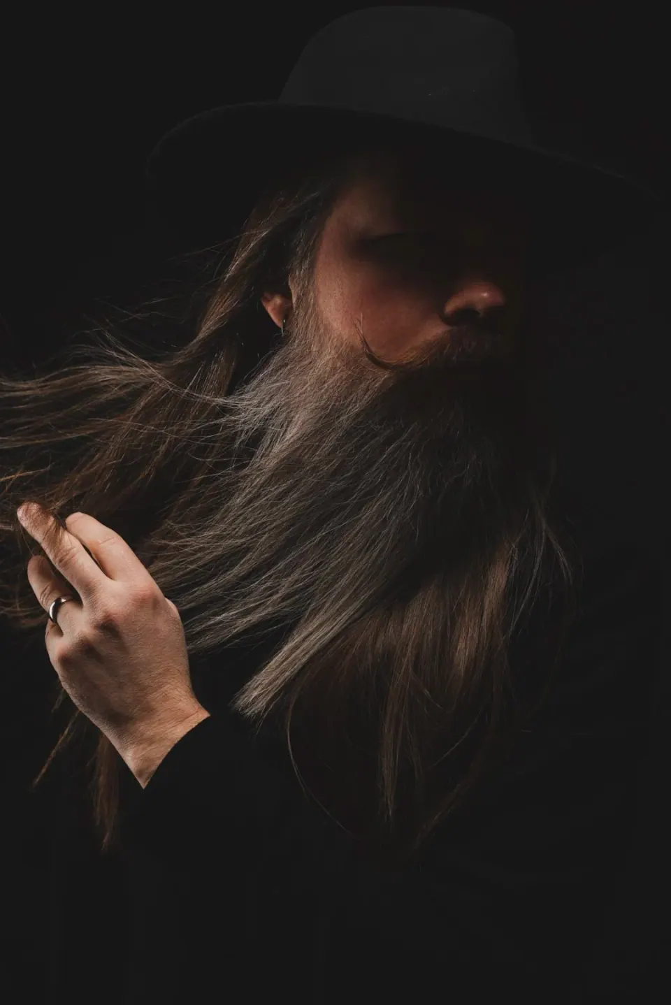Retrato de un hombre con barba y pelo largos, con sombrero negro, sobre fondo negro, con una mano en el hombro.